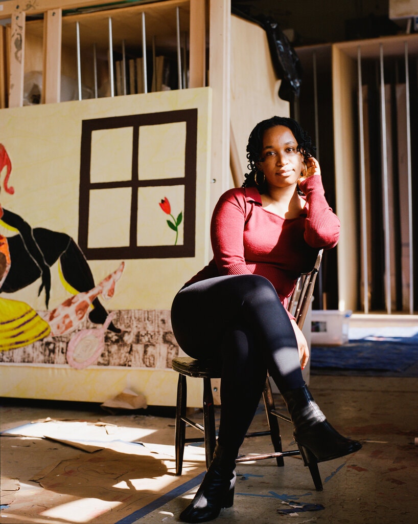 A dark-skinned woman sits in a studio with her legs crossed and body leaning against the back of a chair. Sun filters in through a window, partially illuminating her frame and the floor.