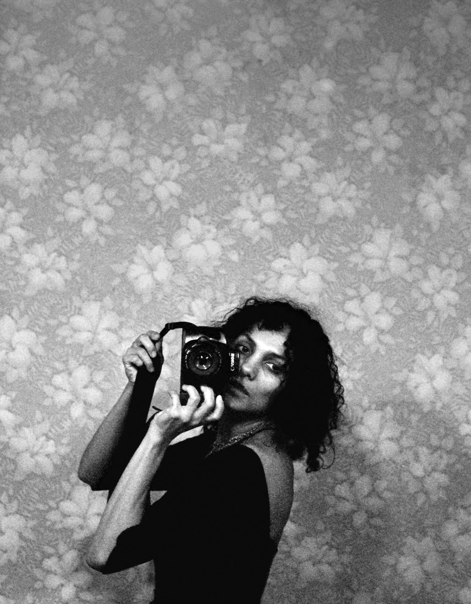 A black-and-white self portrait of a woman standing in front of patterned floral wallpaper. She stands to the side and turns her head forward, holding a camera up. She is visible from the waist up and wears a black leotard.