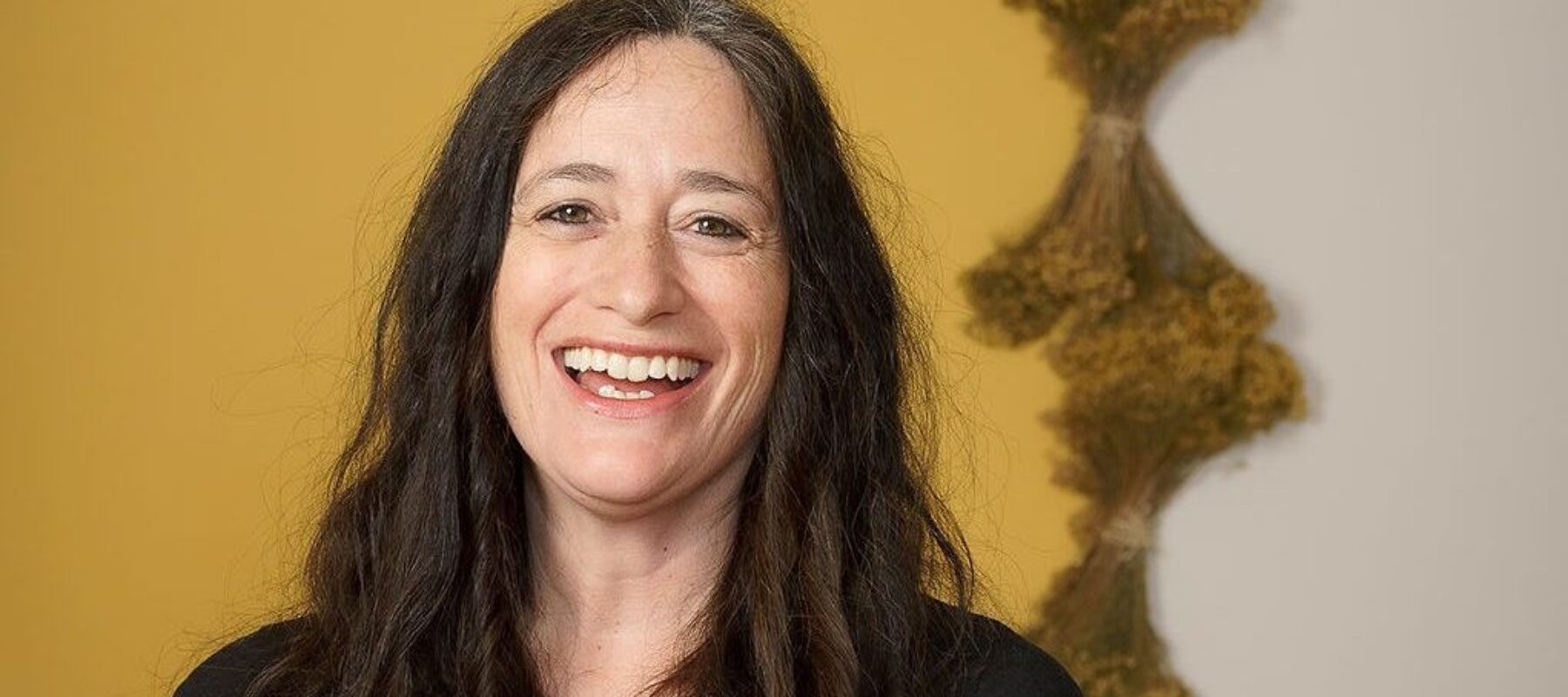 A light-skinned woman with long brown hair smiles happily at the camera. She wears a black t-shirt.