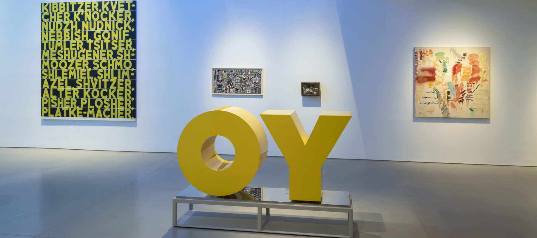In a museum gallery space, a large yellow sculpture of the word "OY" is in the center of the room with four paintings of large and small sizes behind it. The white wall is awash in blue light.