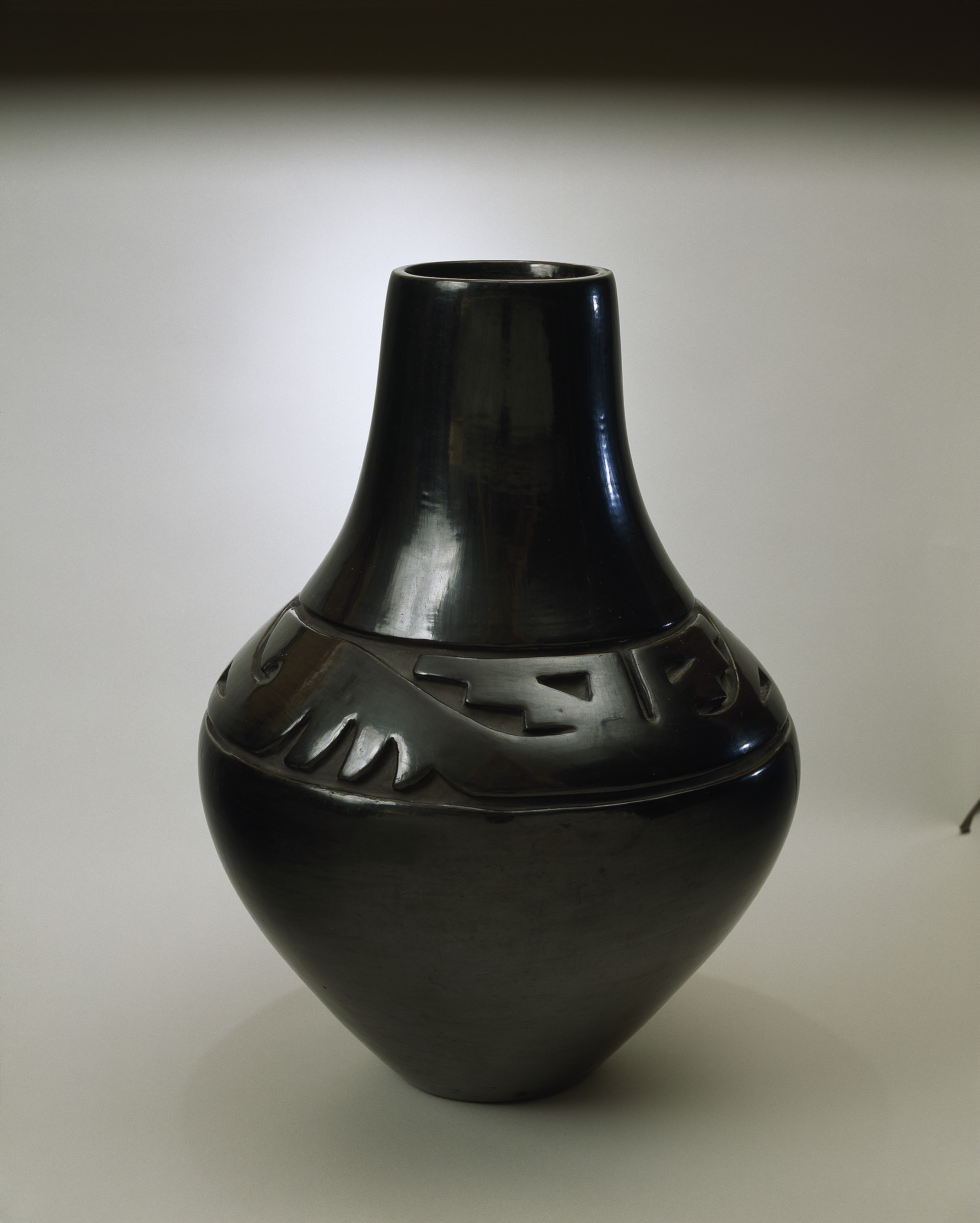 Blackware pottery vessel with tall neck and broad shoulder tapering to a narrow base. The flawless, polished black surface is adorned with deep relief carvings of stylized wings and geometric designs on the shoulder.