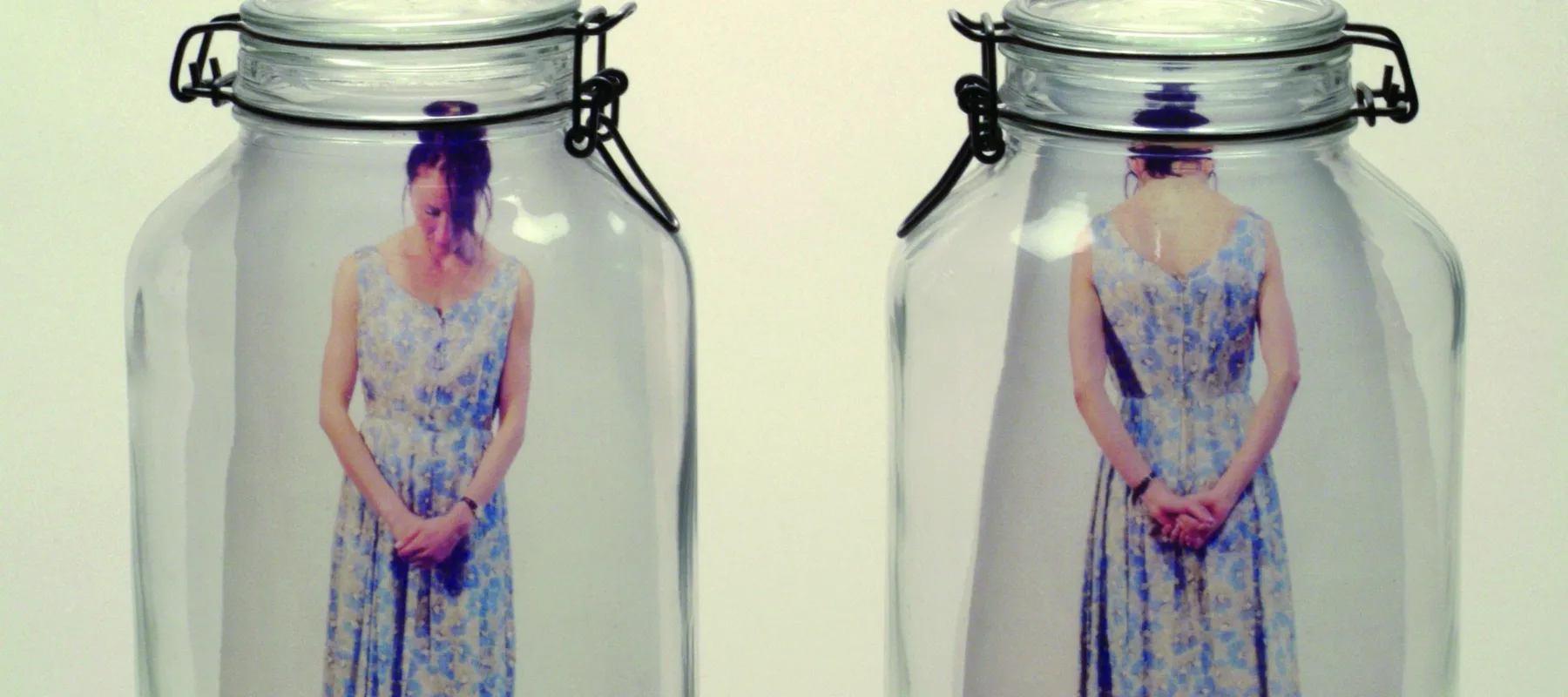 Two clear glass mason jars side by side. In the left jar is a color photograph of a light skinned woman standing with head bent and hands clasped in front of her. In the right jar is the same woman seen from rear with hands clasped behind her.