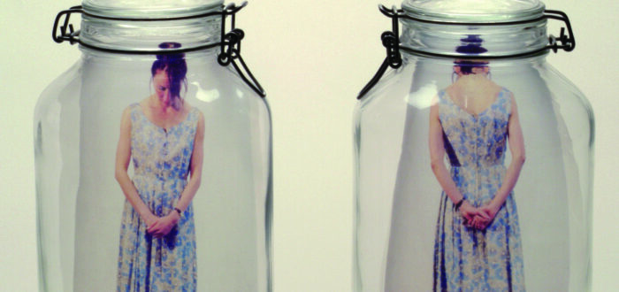 Two clear glass mason jars side by side. In the left jar is a color photograph of a light skinned woman standing with head bent and hands clasped in front of her. In the right jar is the same woman seen from rear with hands clasped behind her.