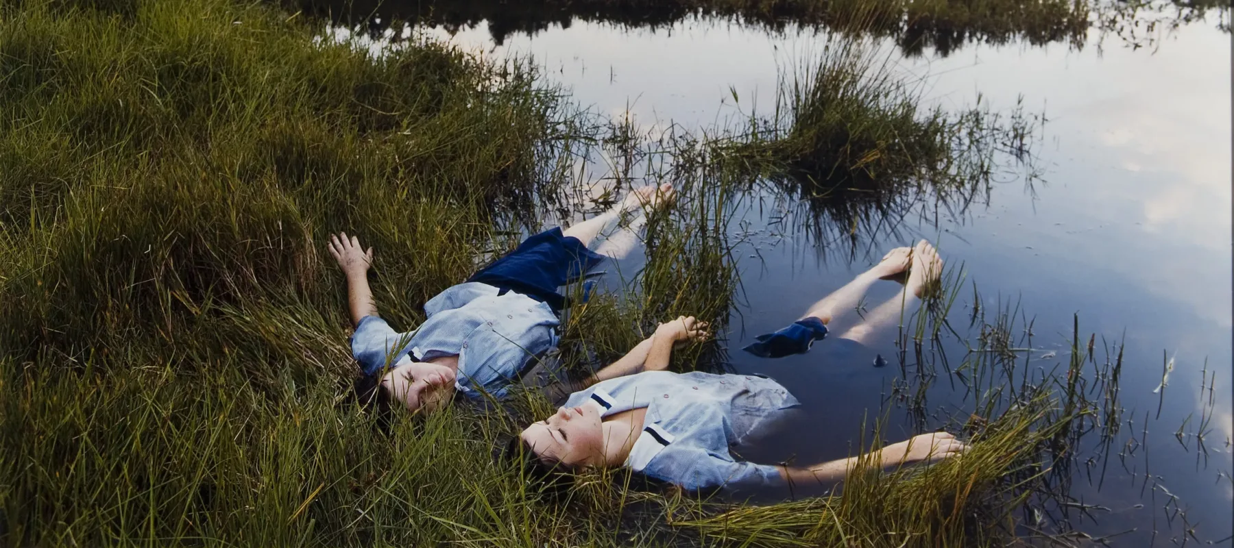 Two adolescent girls with light skin wearing matching blue uniforms float side-by-side on the edge of a grassy pond while holding hands.