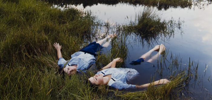 Two adolescent girls with light skin wearing matching blue uniforms float side-by-side on the edge of a grassy pond while holding hands.