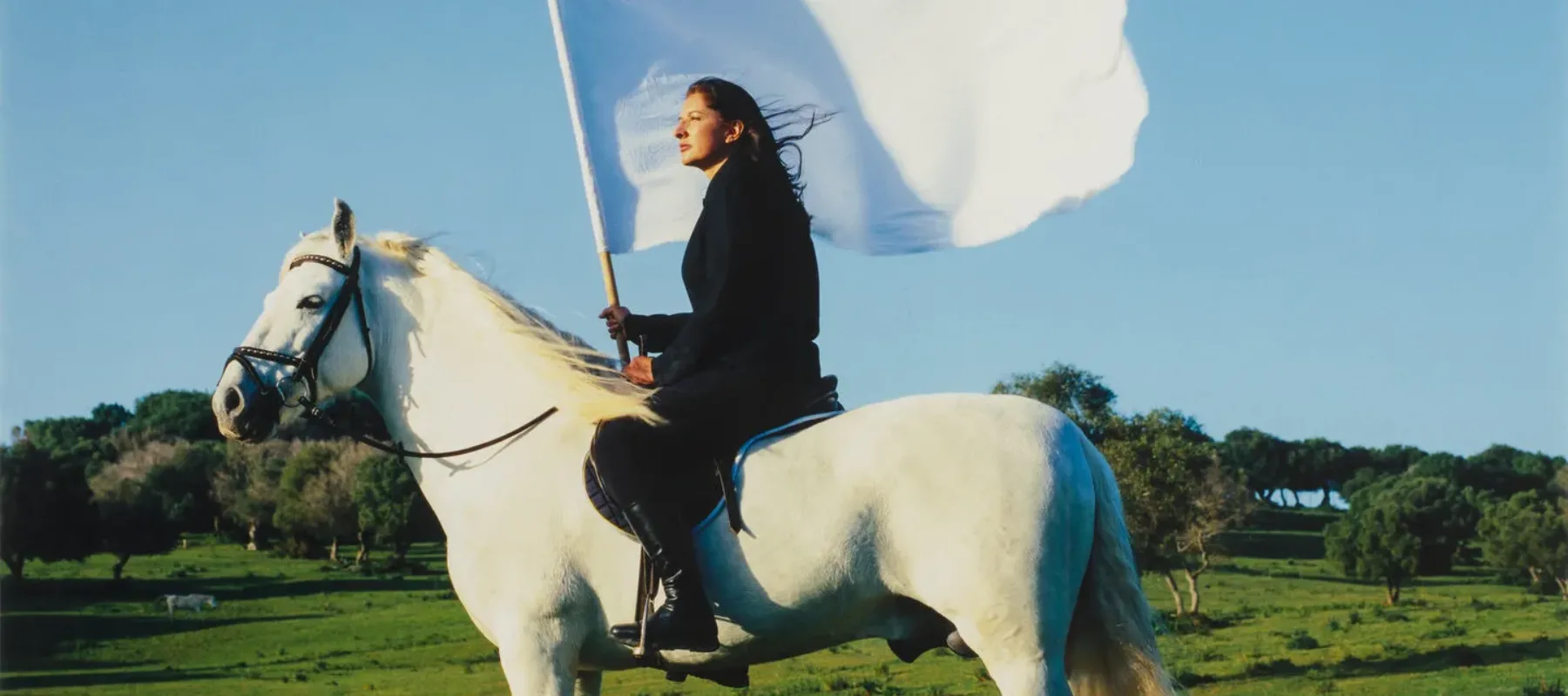 Color photograph of a light skin toned woman in all black on a white horse and carrying a large, white flag. Horse and rider are facing left and are in a rolling green landscape with a bright blue cloudless sky.