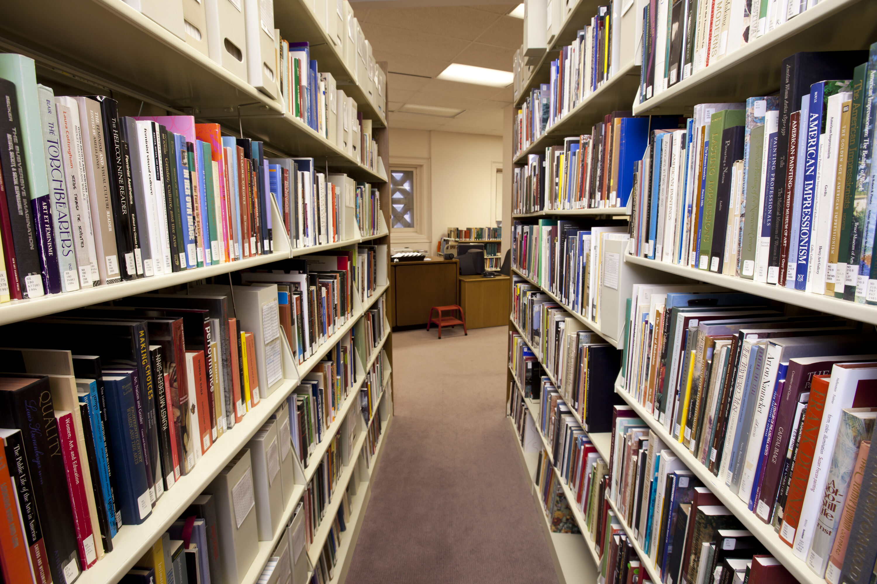 View of an aisle between two tall library bookshelves filled with multi-colored books. At the end of the aisle is a desk with a small orange footstool in front of it and a small bookshelf behind it.