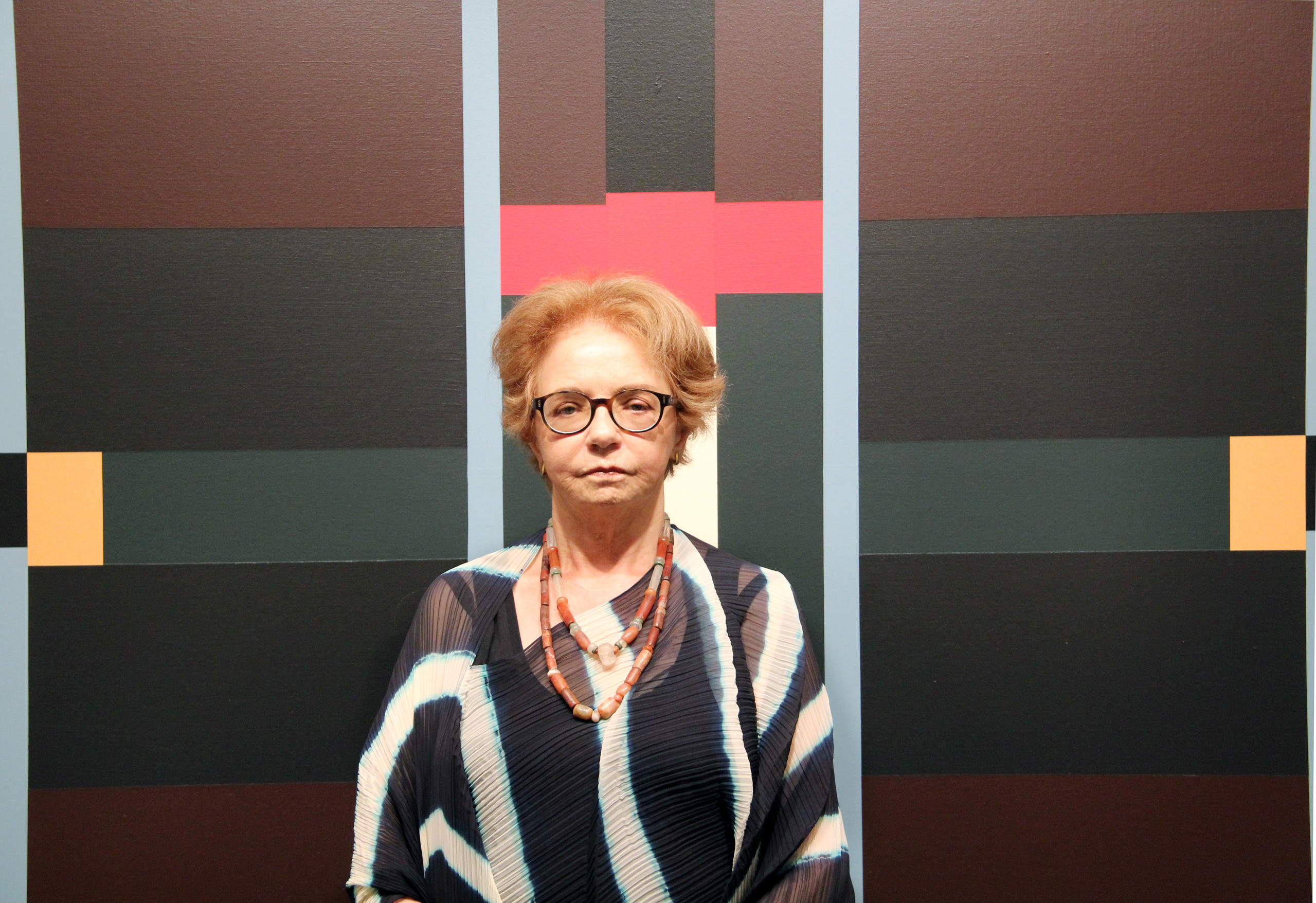 A light-skinned older woman with short, light brown hair stands in front of an abstract painting. She wears glasses, a black and white striped shirt, and two beaded necklaces. The painting behind her depicts black and maroon planes atop which red, teal, peach, and light blue lines and rectangles are rendered.