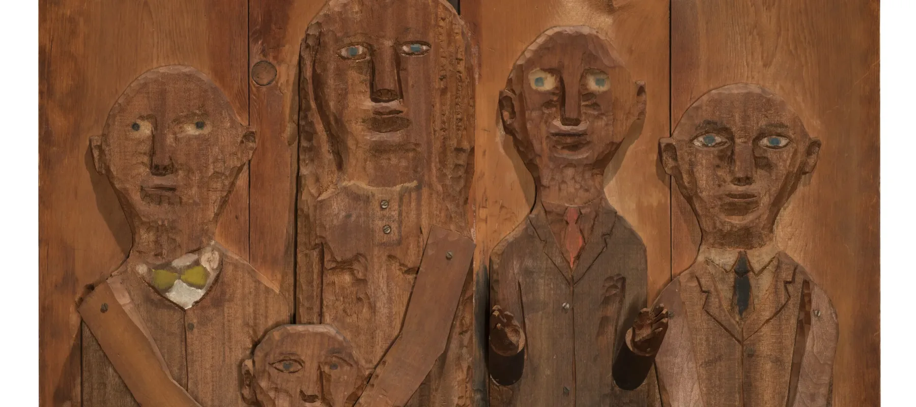 A wooden relief sculpture of four adults and one child. The two figures on the left put their hands on the shoulders of the child in front of them. The arms and hands of all figures are three dimensional and protrude from the work.