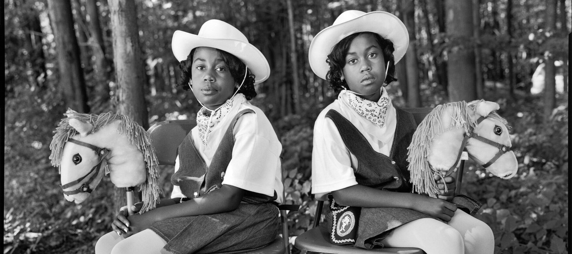 A black-and-white photograph of two dark-skinned young twins sitting in folding chairs against a woodsy background, facing the camera with their bodies angled outward in opposite directions. They wear matching cowgirl outfits, complete with hats, boots, and hobby horses.