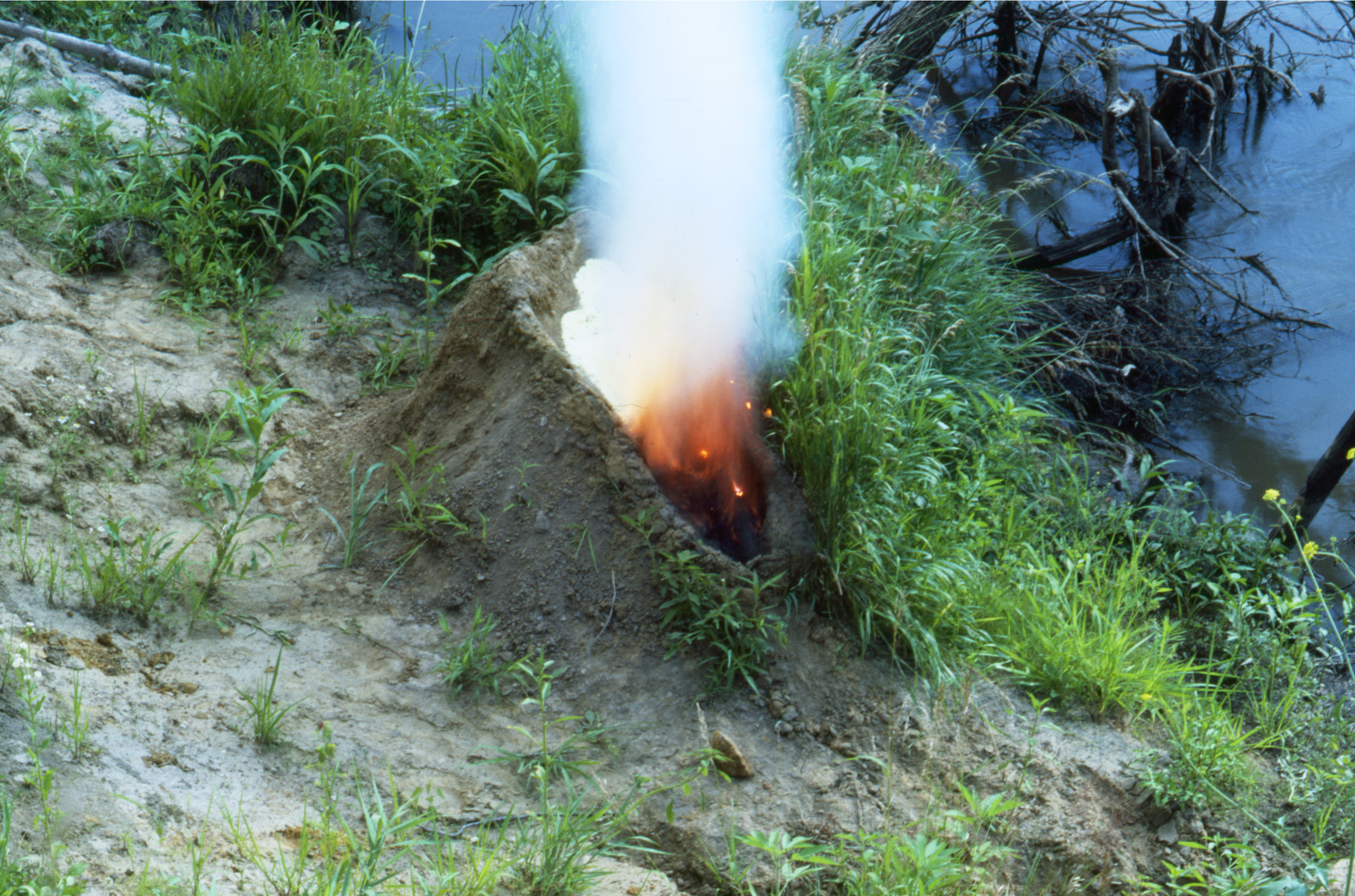 A mound of dirt in a grassy area by water. In the center of the mound is a human-shaped recess full of gunpowder that has been set aflame, exploding up in fire and white smoke, making the dirt mound resemble an erupting volcano.