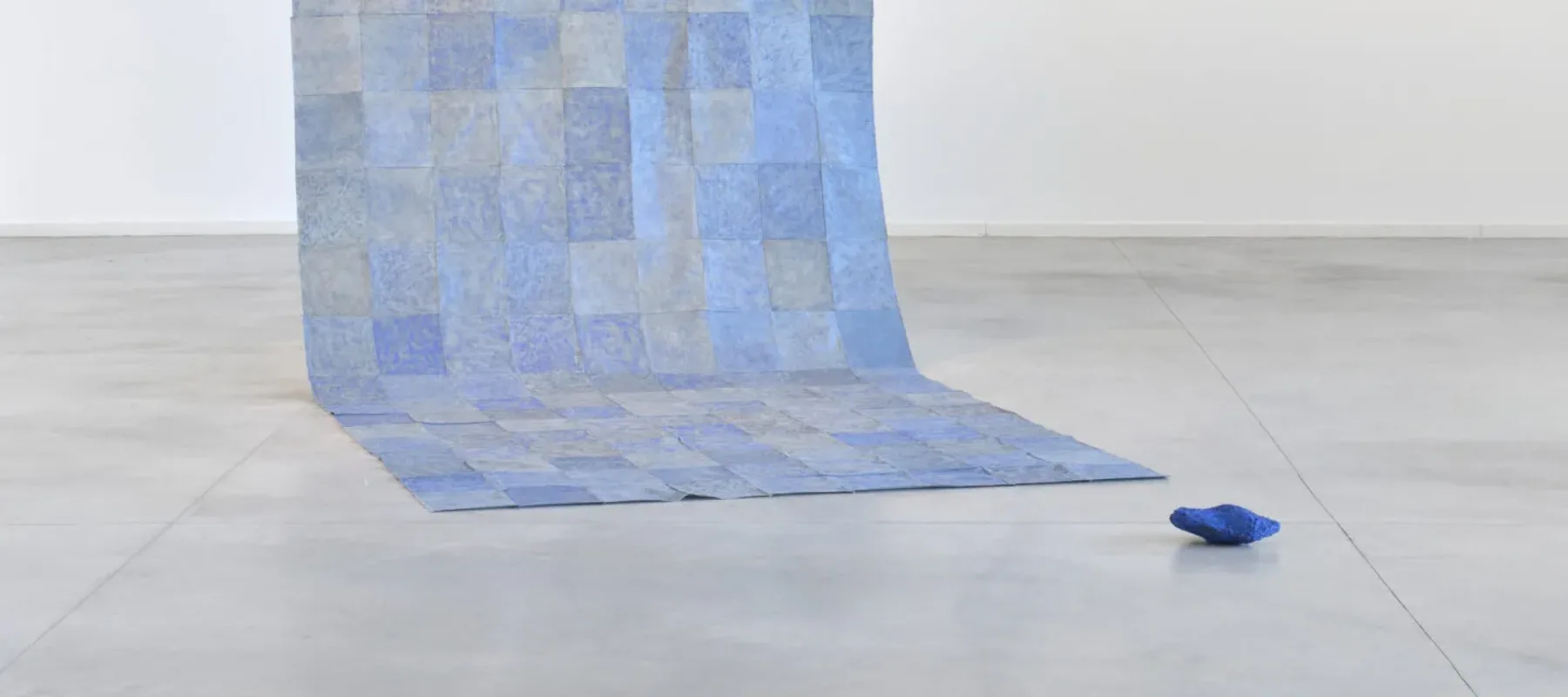 A floor to ceiling contemporary installation of blue square sheets patched together into a long panel that hangs down from two wires and partly rests flat on the floor. A dark blue object lays nearby.