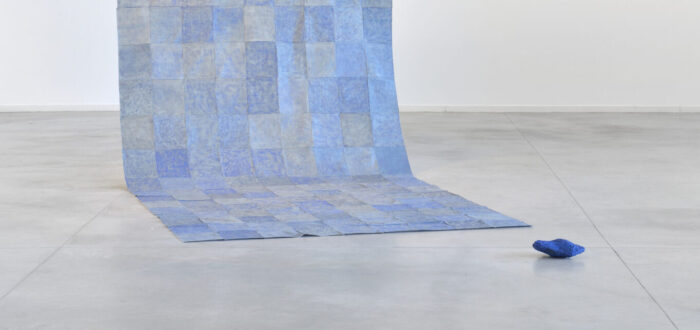 A floor to ceiling contemporary installation of blue square sheets patched together into a long panel that hangs down from two wires and partly rests flat on the floor. A dark blue object lays nearby.