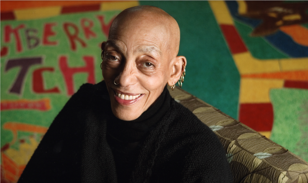 A dark-skinned older woman smiles with warmth at the camera. She is bald and wears five hoop earrings up the length of her right ear and a small hoop earring in her left nostril. Behind her is a colorful abstract artwork, seemingly made from felt, in colors of yellow, red, and green.