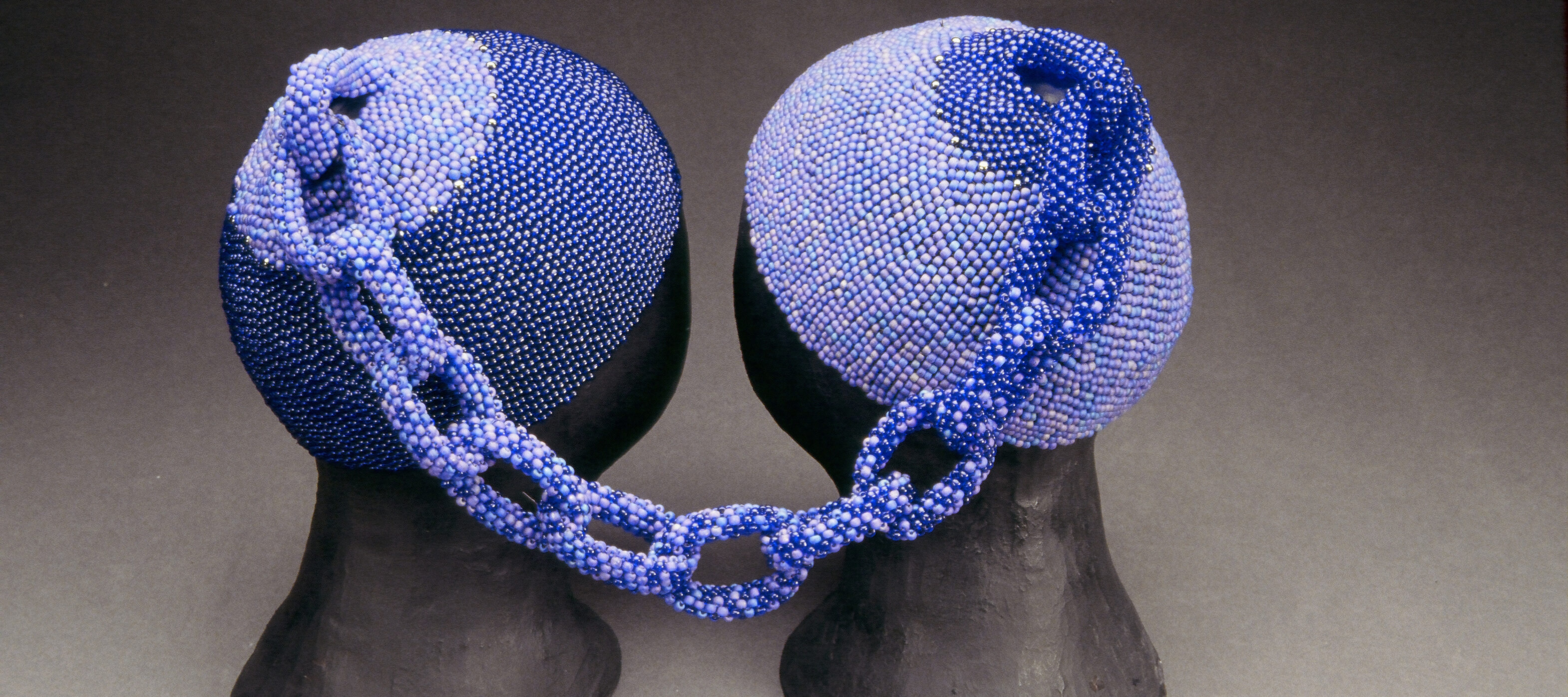Two head caps made of small, blue glass beads rest on two black mannequin heads. The two caps are connected at the tops by a beaded chain. The left cap is made of darker blue beads and the right cap is made of lighter blue beads. The chain combines both shades.