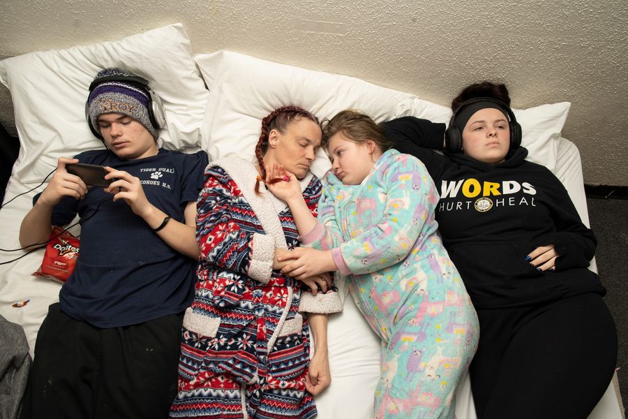 A photograph shot from above shows four light-skinned people laying side-by-side in a bed. On the left, a teenage boy wears a beanie hat, headphones, and plays on his phone. Next to him, a middle-aged woman wears a fluffy patterned robe, french braid pig tails, and lies with her eyes closed facing a younger girl to her right. The girl holds the woman's hand and places her other hand on the woman's face while looking at her; she wears a onesie. Next to her a teenage girl stares at the ceiling with headphones on, wearing a black sweatshirt and black pants. Her shirt says "Words Hurt Heal."