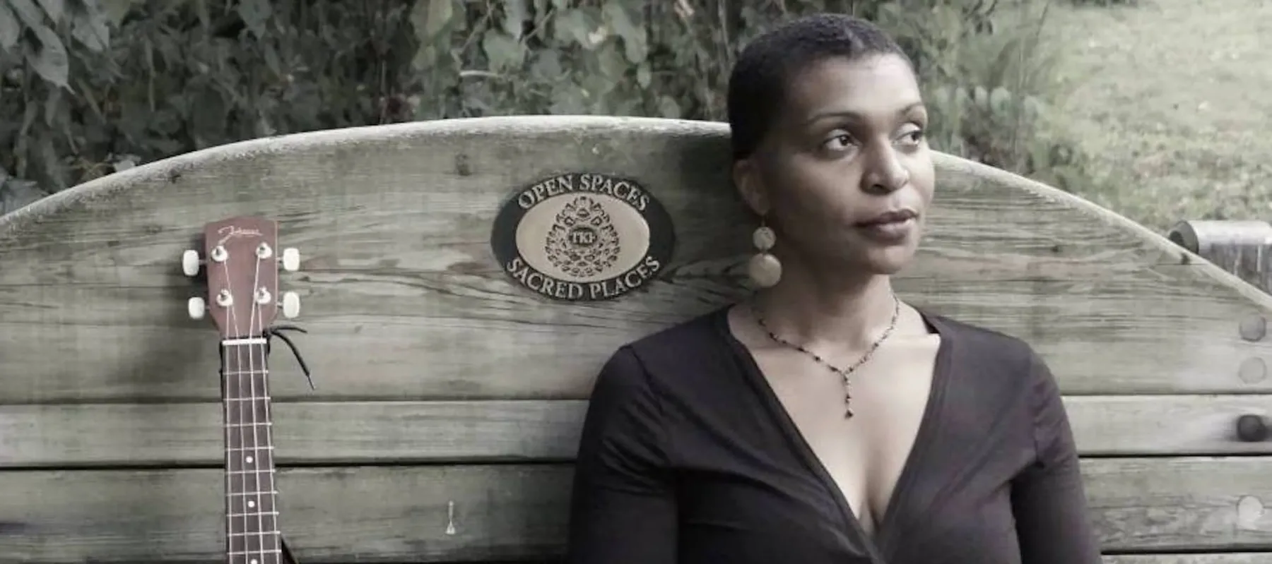 Courtney Dowe sits on a wooden bench with a high back with a standing brown guitar on her right. Her legs are crossed and her arms are crossed on her lap. She wears a black top, a patterned skirt, earrings, and a necklace, and she has very short hair. Behind her is lush greenery.