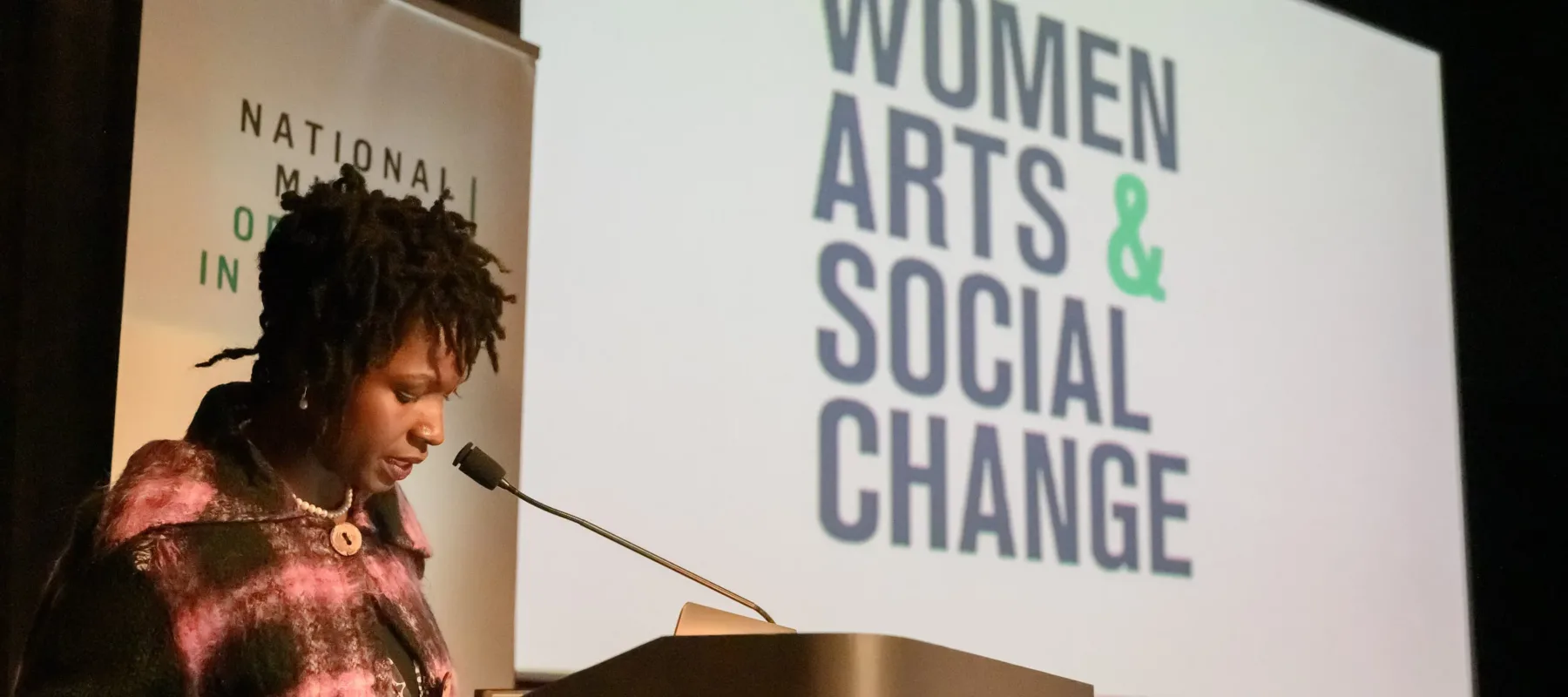 A dark-skinned adult woman stands at a podium at the National Museum of Women in the Arts to speak. She has short dreadlocks, jewelry, and a jacket with a large purple and black plaid print on it. Behind her is a screen with the words 'Women, Arts, & Social Change.'