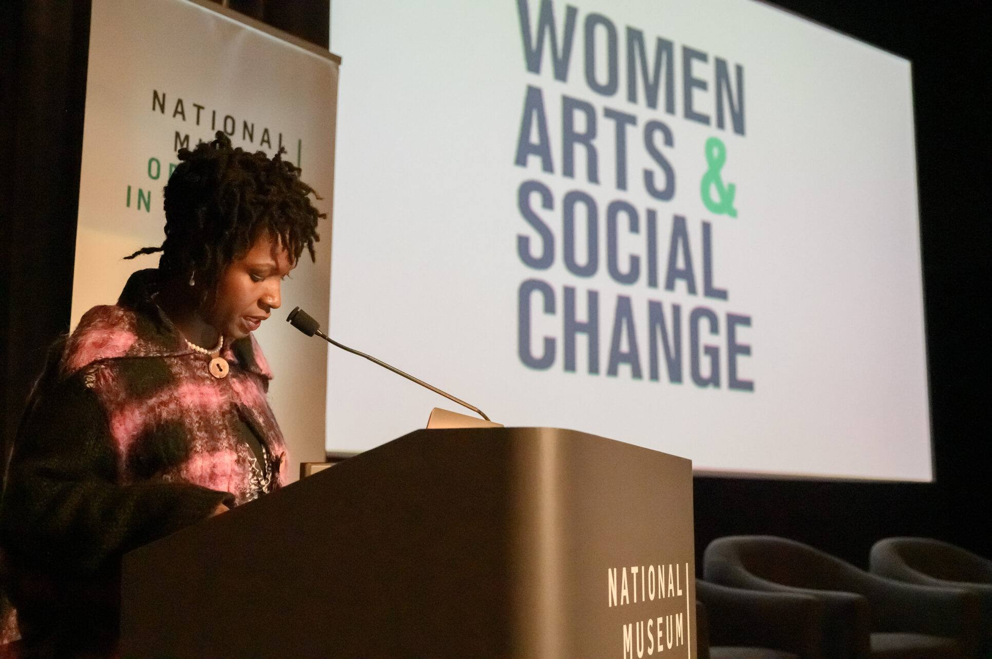 A dark-skinned adult woman stands at a podium at the National Museum of Women in the Arts to speak. She has short dreadlocks, jewelry, and a jacket with a large purple and black plaid print on it. Behind her is a screen with the words 'Women, Arts, & Social Change.'