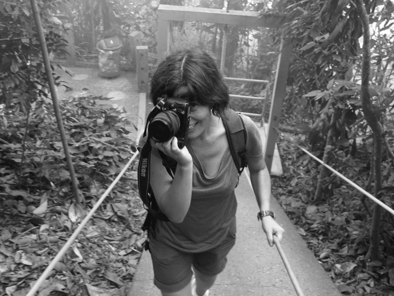A black-and-white photograph of a light-skinned woman in shorts and a t-shirt, walking up a sidewalk in a jungle or tropical setting. She holds a thin railing on her right and with her left hand holds a Nikon SLR camera up to her eye, smiling, as if to take a photo of her photographer.