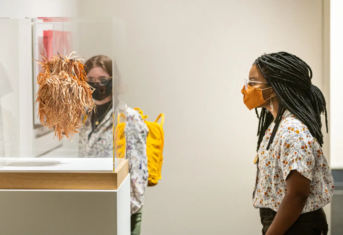 Young woman with long, thin braids, wearing glasses and an orange face mask, looks at a small hanging sculpture made of thinly cut strips of pale orange paper.