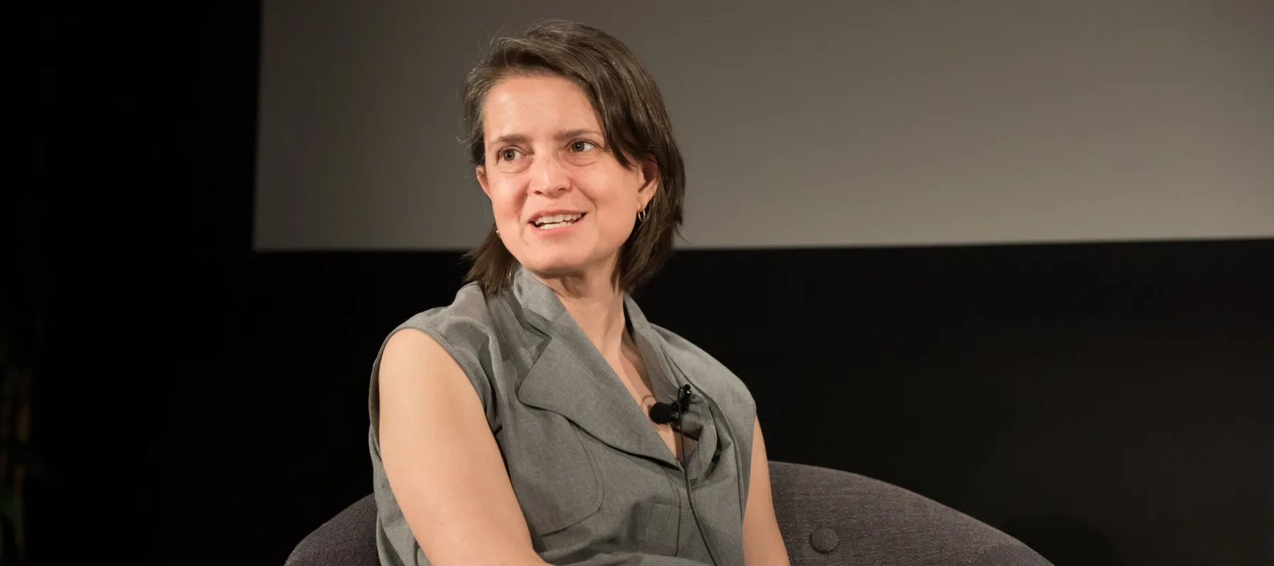 A light-skinned woman with short, chin-length straight brown hair sits on a plush grey chair on what looks like s stage. She looks out to the left and is seemingly mid-sentence. Her legs are crossed and she wears a long sleeveless grey shirt over black pants.