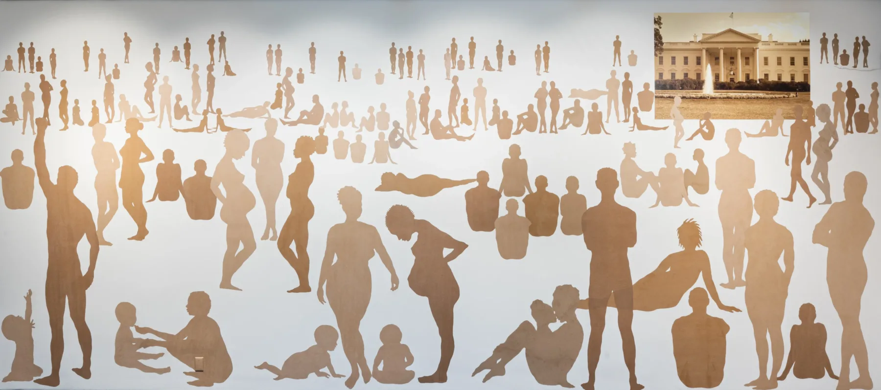 Over 100 silhouetted figures of varying ages, shapes, poses, and skin tones on a large white wall. In the top-right corner is a rectangular picture of the White House.