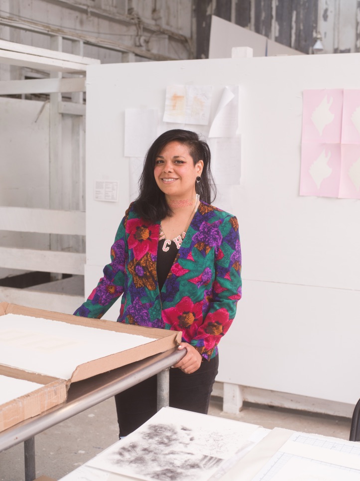 A medium-skinned woman with black straight hair that is long on the left side and shaved on the right stands smiling in an industrial space. She wears a bright floral blazer in pink, green, and purple.