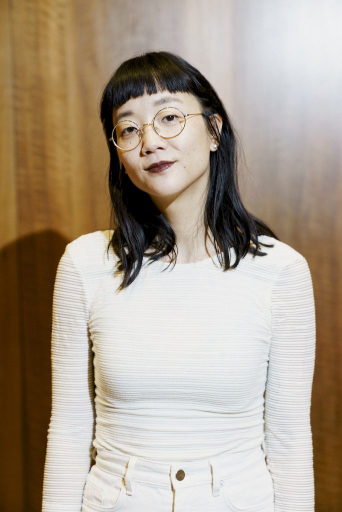 A casual portrait photograph of a light-skinned Asian woman wearing a tight white long sleeved shirt and white jeans. She smiles slightly at the camera. She wears her black hair down at shoulder-length and circular, thin-rimmed classes.