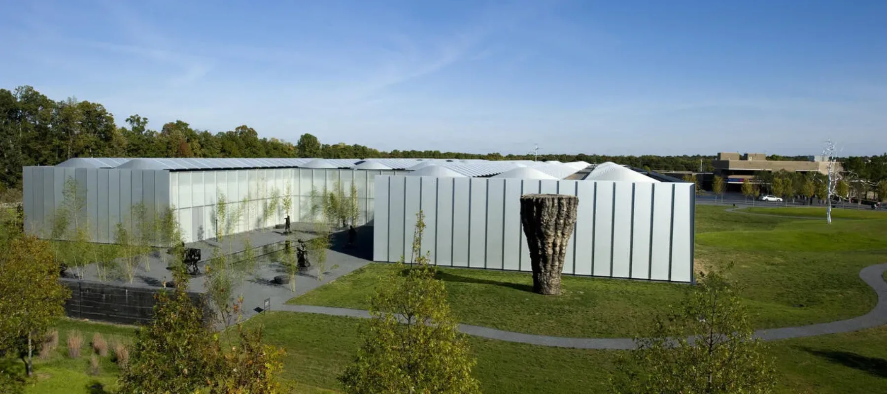 An aerial shot of a landscape featuring a large white modern building, a large wooden sculptural form, and blue skies with clouds and and an expansive green lawn.