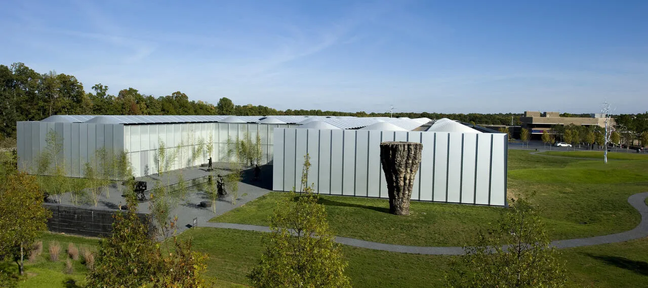 An aerial shot of a landscape featuring a large white modern building, a large wooden sculptural form, and blue skies with clouds and and an expansive green lawn.