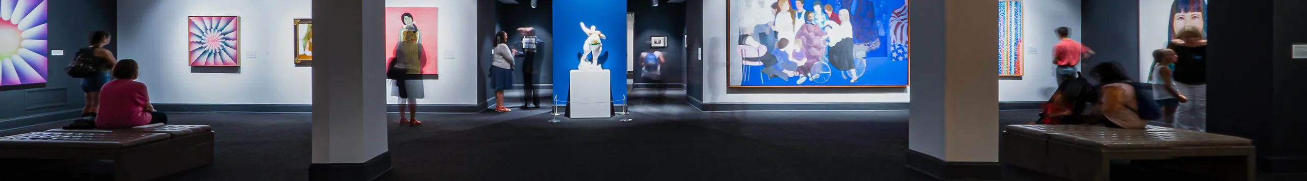 Wide angle view showing a full gallery. The floor is a dark gray carpet and there are two columns, through which large scale works in vibrant colors are visible.