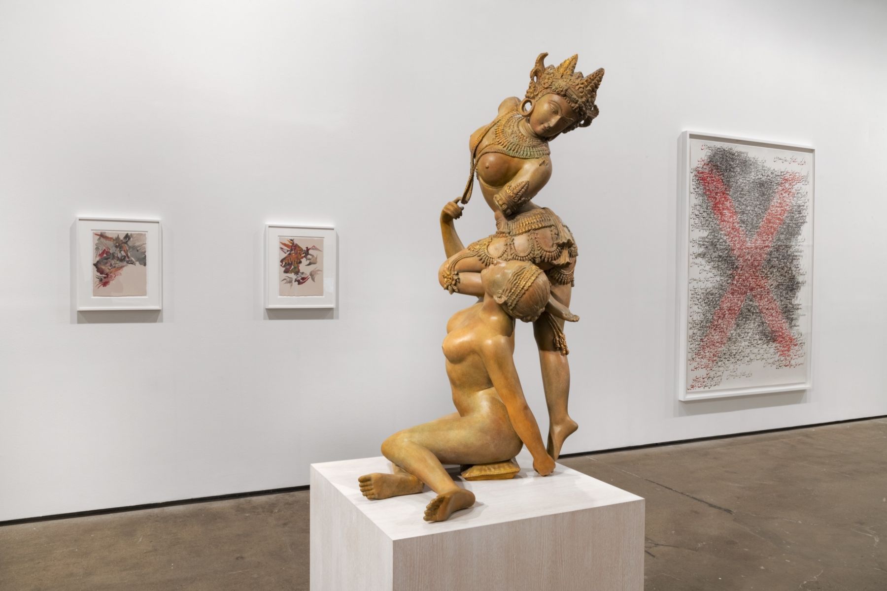 A light brown sculpture of two women, one appearing as a goddess from Southeast Asia, is on display in the middle of a contemporary gallery space. On the wall behind it three abstract paintings hang.