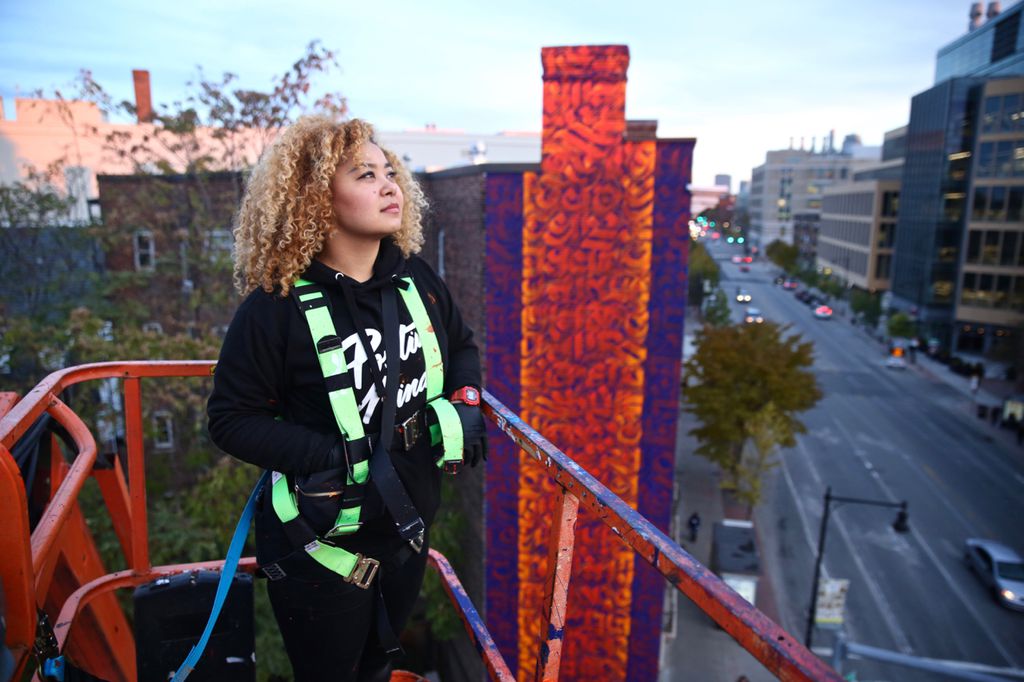 A medium-skinned woman with light brown curly hair stands in a construction box, elevated over city streets. She wears a safety harness and looks off into the distance. Behind her a vibrant mural is rendered on the side of a building in bright orange and purple. It is a pattern, but the exact makeup is obscured.