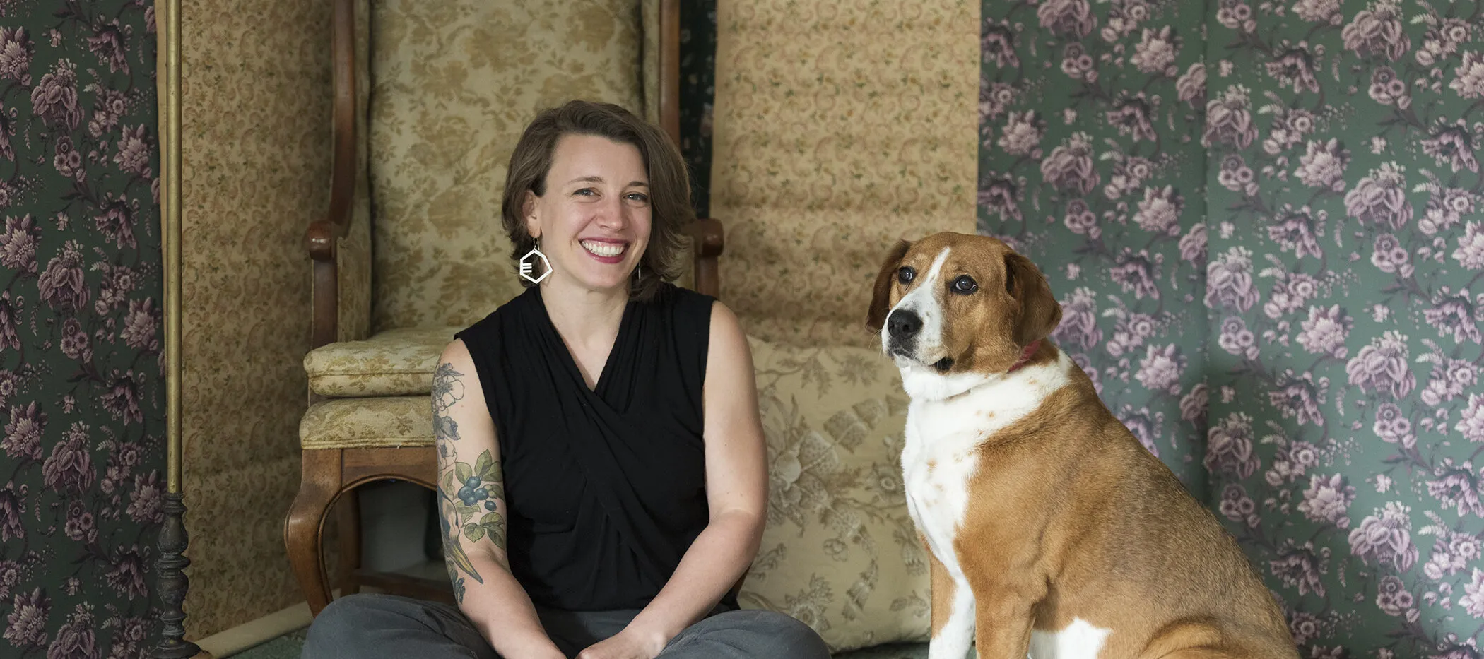 A light-skinned woman with shoulder-length brown hair smiles at the camera. She wears octagonal earrings and a sleeveless black shirt that reveals one arm full of flower tattoos. Sitting next to her is a brown and white dog, looking off into the distance. They are both positioned in front of floral patterned wallpaper and an antique chair, also covered in a floral pattern.