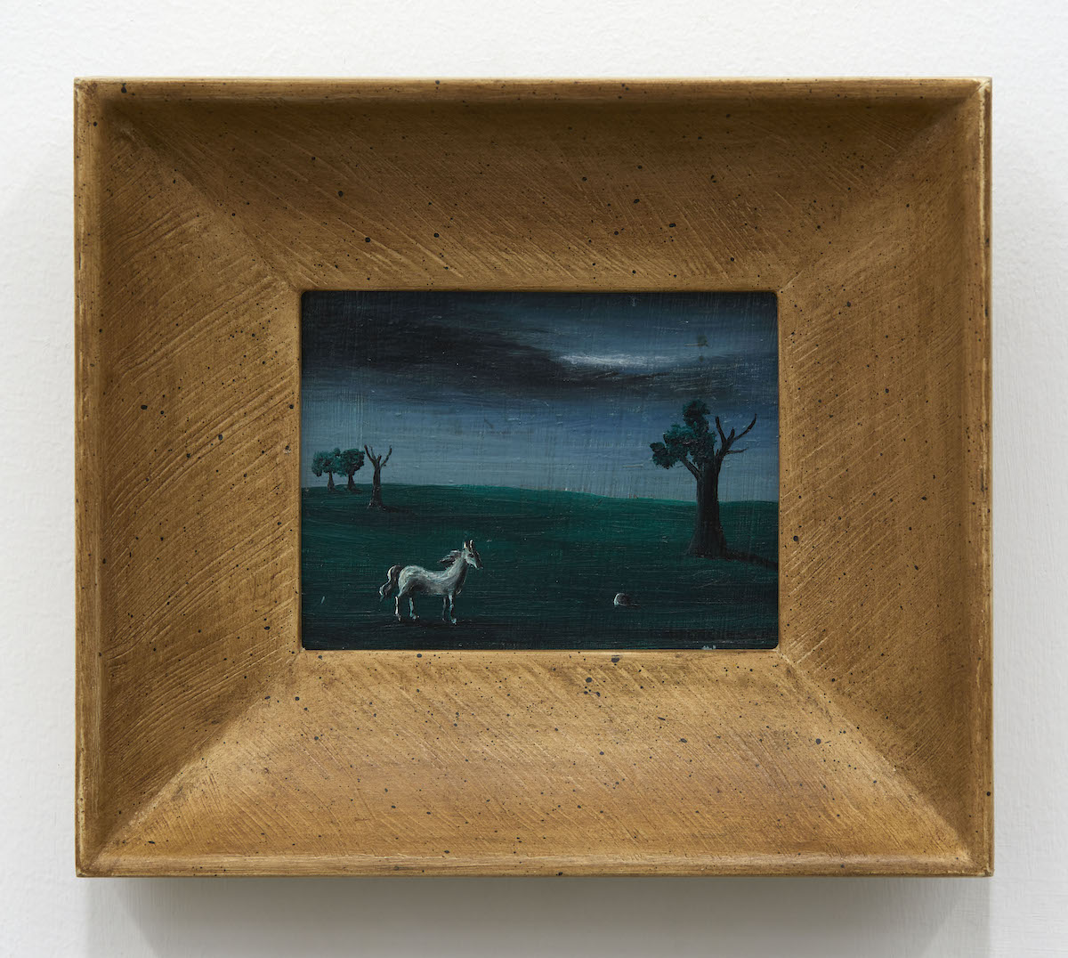 Inside a thick gold frame is a tiny Surrealist painting depicting a dark, moody sky and landscape. Three trees are in the background and one larger one is in the foreground atop dark green grass. Half the tree is dead, the other with leaves. One lone white horse stands towards the bottom of the frame.