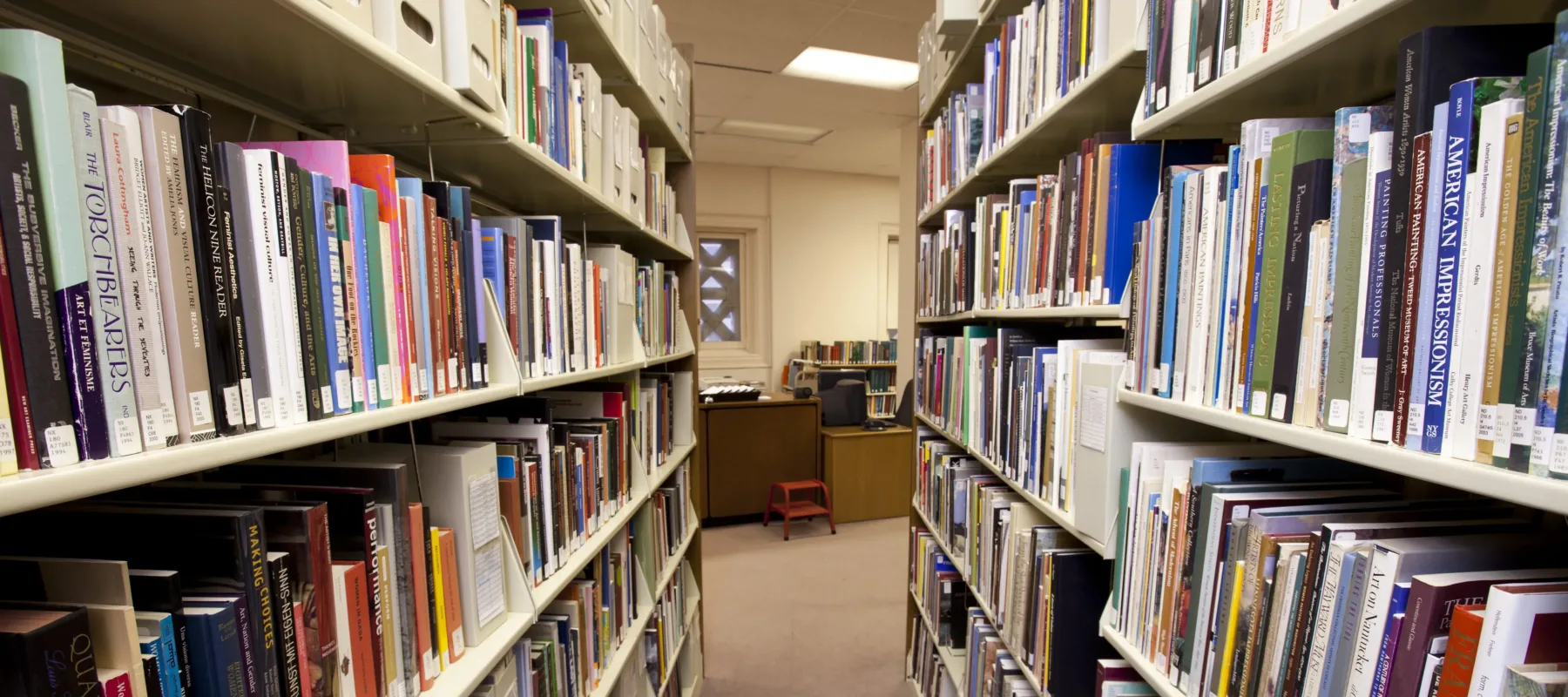 View of an aisle between two tall library bookshelves filled with multi-colored books. At the end of the aisle is a desk with a small orange footstool in front of it and a small bookshelf behind it.