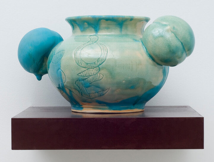 A round, ceramic vessel glazed unevenly in glossy celadon has a bulging body incised with abstract shapes. Matte turquoise glaze dribbling down from the rim obscures portions of the pale green and fully coats one of two peach-shaped orbs projecting from either side of the neck.