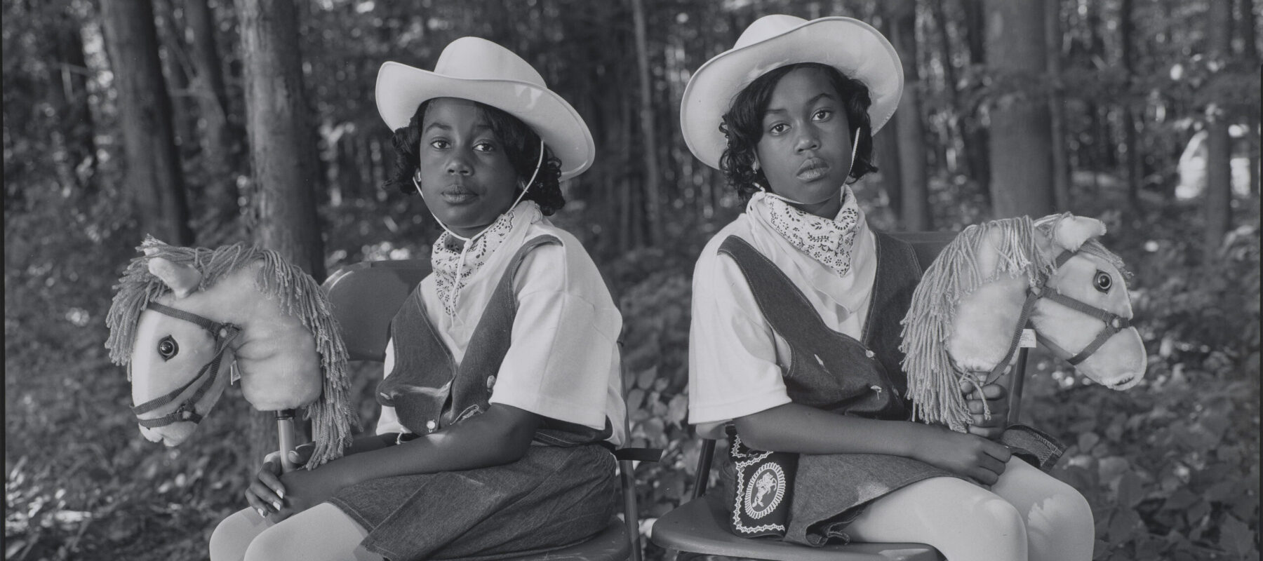 A black-and-white photograph of two dark-skinned young twins sitting in folding chairs against a woodsy background, facing the camera with their bodies angled outward in opposite directions. They wear matching cowgirl outfits, complete with hats, boots, and hobby horses.