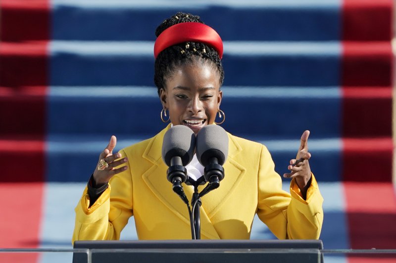 A young African American woman stands at a podium in front of two large microphones. She is mid-sentence, gesturing outwards with both arms. Her hair is braided and pulled back from her face; she wears a ruby read silk headband like a crown, a bright yellow pea coat, and small gold hoop earrings.