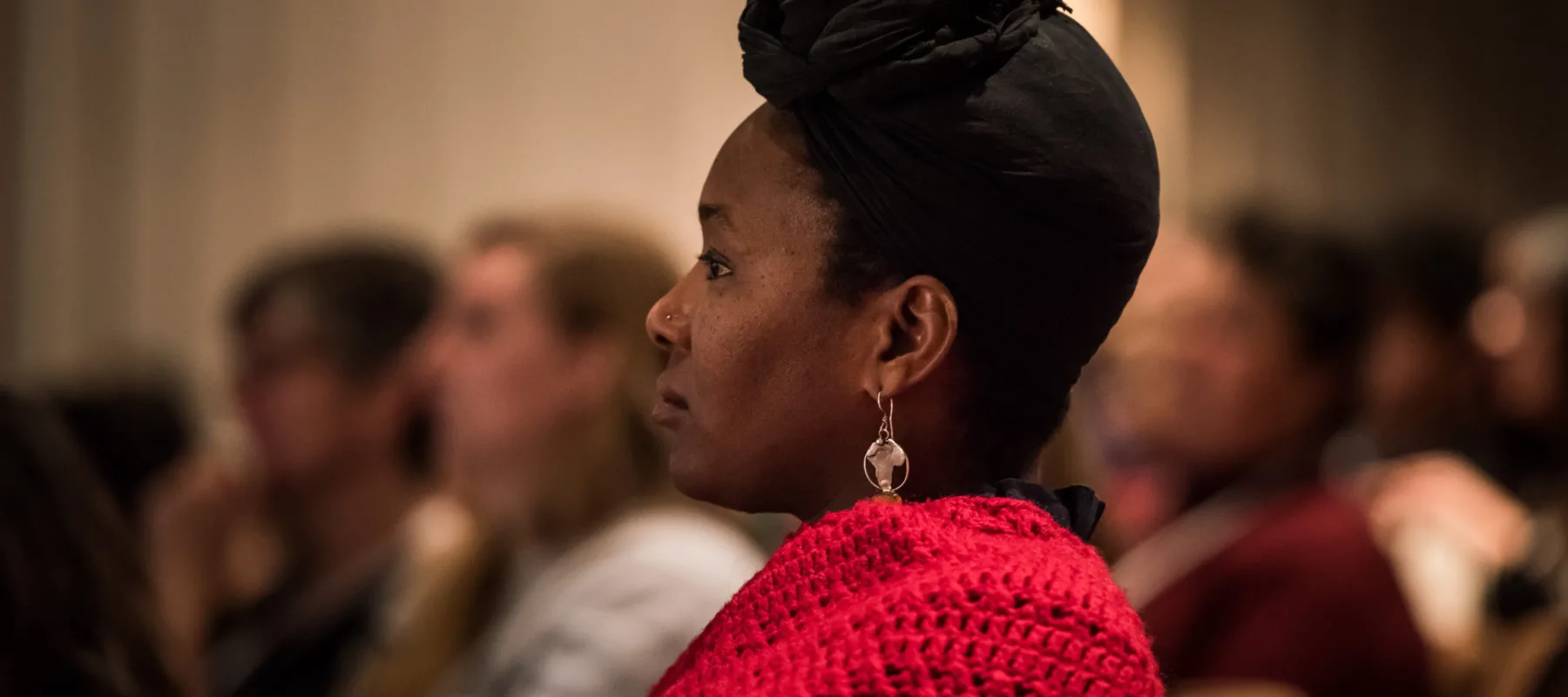 A dark-skinned adult sitting in profile, seen from the shoulders up. They wear a red sweater, gold earrings, and their hair tied up in a black headscarf. They sit in an auditorium, with blurry figures filling the seats in the background.