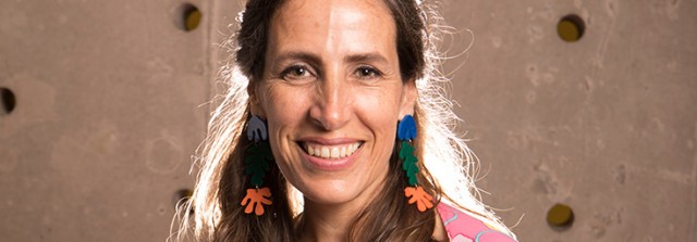 A portrait photograph of a woman with a light skin tone and long, blonde hair wearing colorful earrings in organic shapes. She is smiling towards the camera.