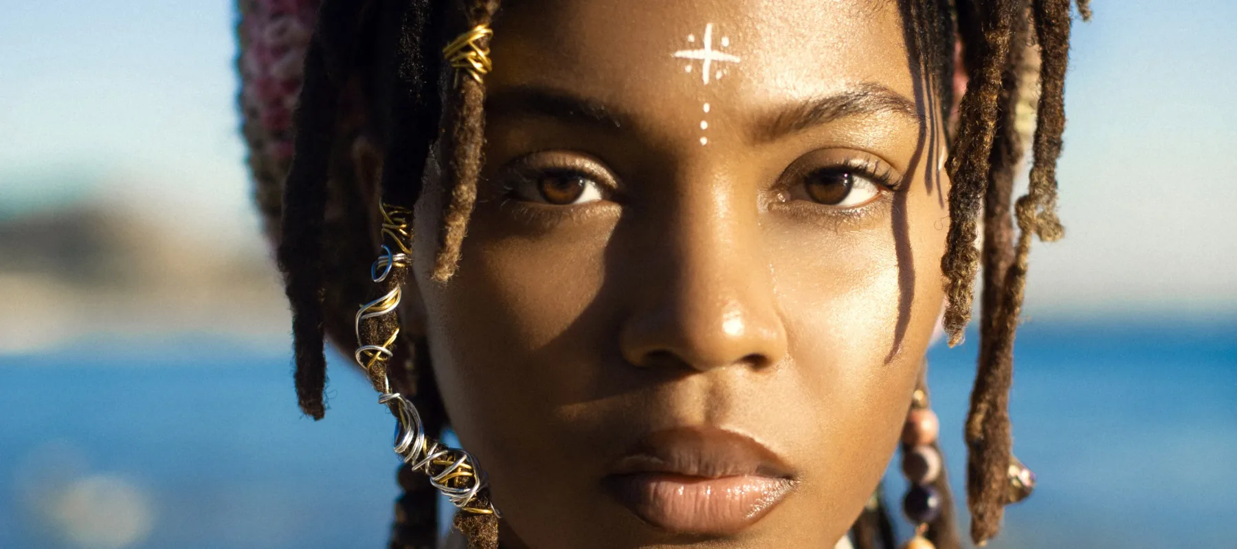 Headshot of MovaKween, a medium-dark skinned adult woman gazing at the viewer. She has brown eyes and thin dreadlocs adorned with wires and beads. A white cross and dots are painted on the center of her brow and behind her is a blurred, vibrant ocean landscape.