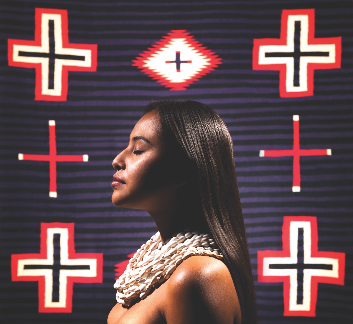 A portrait photograph of a medium-skinned Indigenous woman standing in profile and facing the left. Her eyes are closed and light shines on her face and chest, which is adorned only in a chunky white necklace collar of small shell beads. Behind her, native cross symbols are painted in red, white, and dark blue atop dark blue and black stripes.