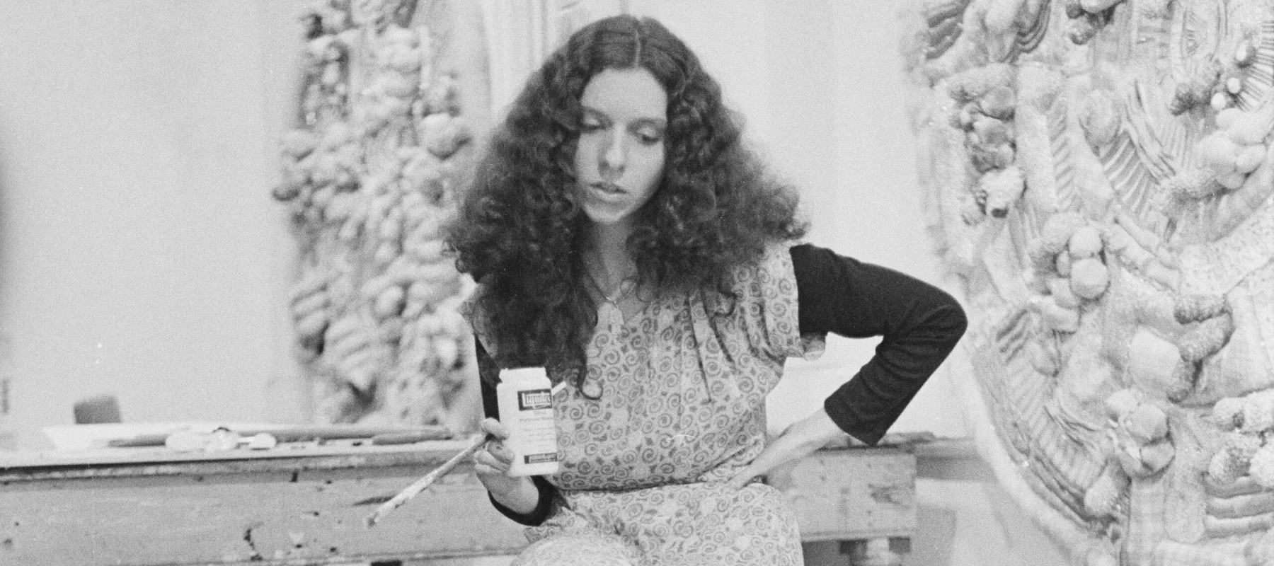 A black-and-white photo of a light-skinned woman sitting on a stool in an artist's studio, looking down at a large circular fabric/textile work. She holds a bottle of gesso and a paint brush.