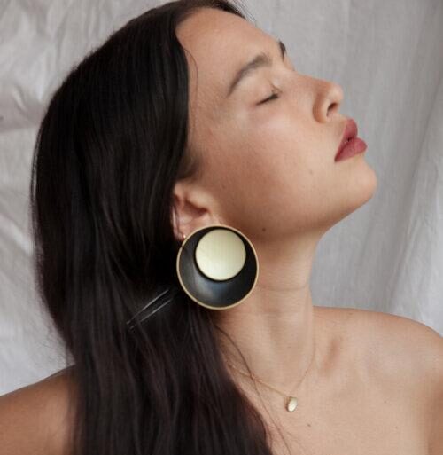 A light-skinned woman with long straight dark-brown hair is captured in profile, her eyes closed and face tilted upward. She wears a large round earring, which is prominently displayed. It is a black ceramic circle filled with another smaller white circle.