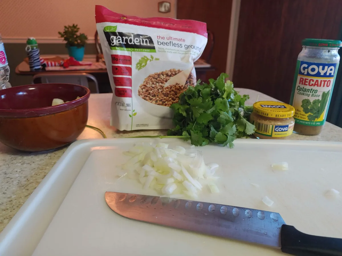 A kitchen scene showing a white cutting board and sharp silver knife placed down next to chopped white onion. On the counter behind it is a plastic package that reads" Gardein--the iltimate beefless ground meat"; a bunch of fresh cilantro, and two glass Goya bottles, one of minced garlic, the other of a cilantro cooking base.