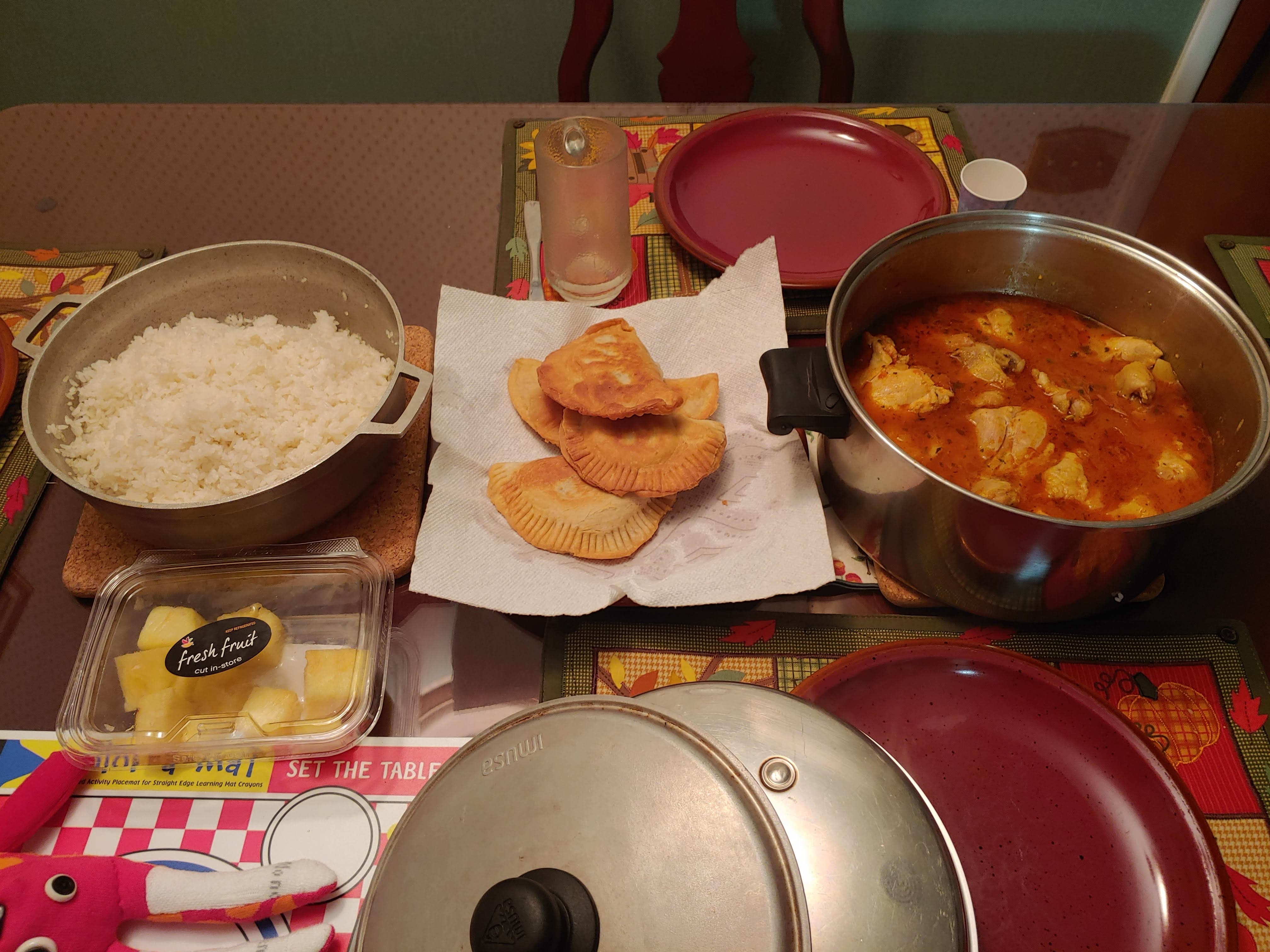 An overhead photo of a dining table that holds one large pot of white rice, four fried beef/flour patties atop a paper towel, a stainless steel pot of chicken in a red sauce, and a plastic container of pineapple. Dark red ceramic plates sit atop a fall/harvest scene on cloth placemats.