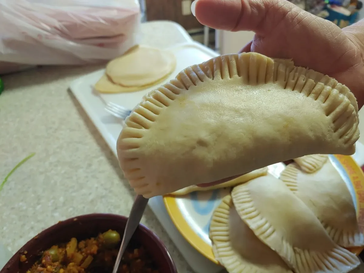 A close-up photograph of a freshly made dough patty held up to the camera. The dough is light colored and the edges have been pinched with fork prongs. Below this patty are others resting on a late and next to them is a bowl of filling, including olives, ground "beef", and onion.