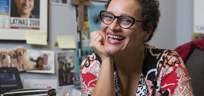 A olive-skinned woman smiles brightly, seemingly mid-laugh, as she sits at an office desk, her right hand placed beneath her chin. Her brown curly hair is worn in a cropped pixie cut and she wears cat-eye glasses. In front of her is a closed Apple laptop; the wall behind her holds two framed certificates or degrees, a framed newspaper, a wooden cross, and other pasted photos.